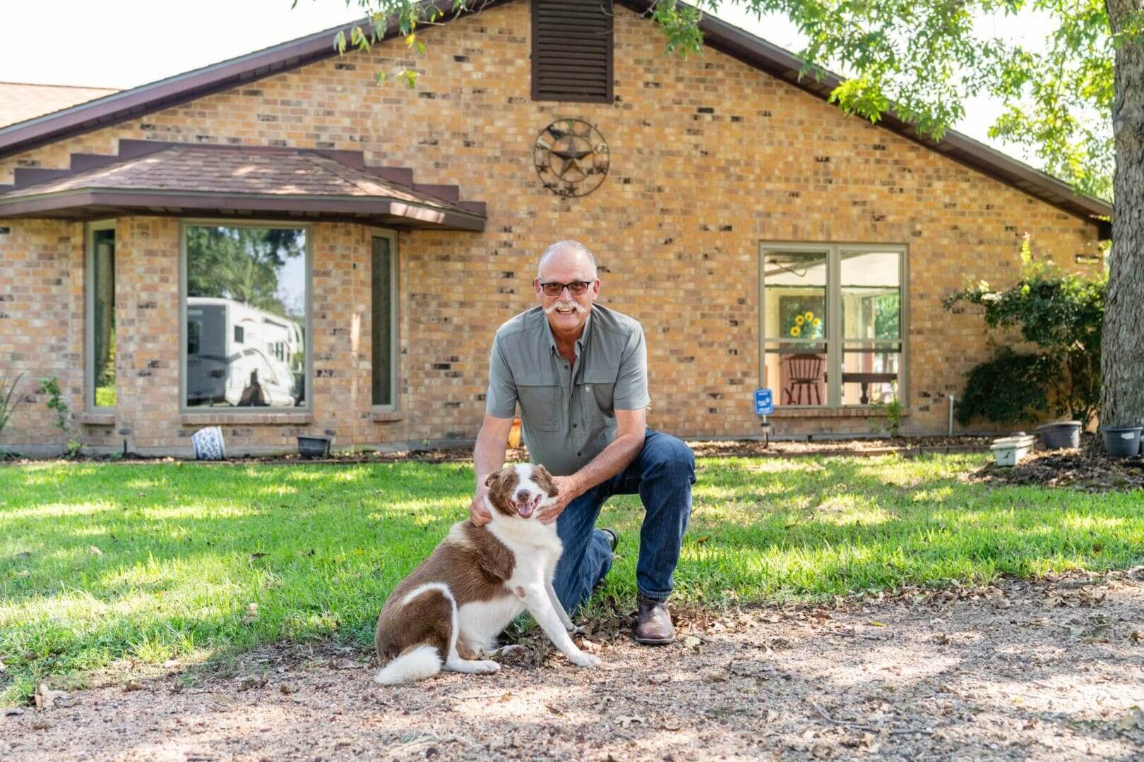 Man with sunglasses kneeling beside a dog outside a brick house.