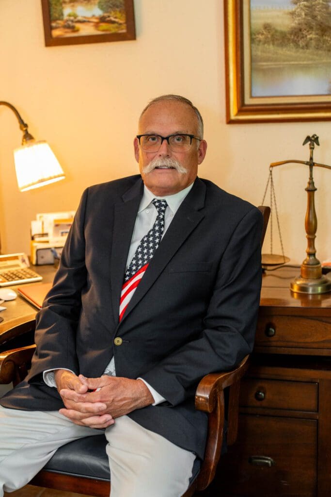 Elderly man in a suit sitting at a wooden desk with a lamp and scales of justice.